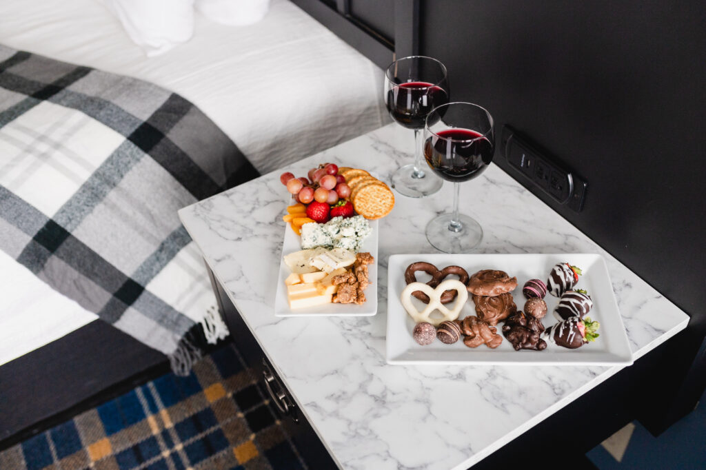 Wine and cheese plate with plate of chocolates on marble table next to a bed with black and grey blanket draped over bed.