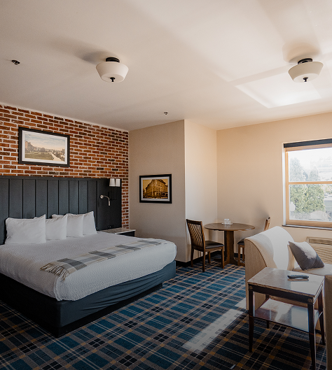 Hotel room with a queen bed, brick wall, plaid carpet, small table with two chairs, and a window.