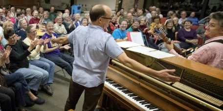A man stands at a piano facing a seated audience, arms spread wide as if leading a singalong.