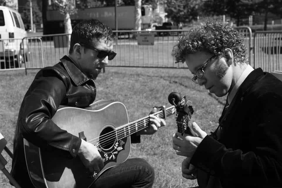 Two men in an outdoor, fenced area; one plays an acoustic guitar while the other looks on closely. Black and white.