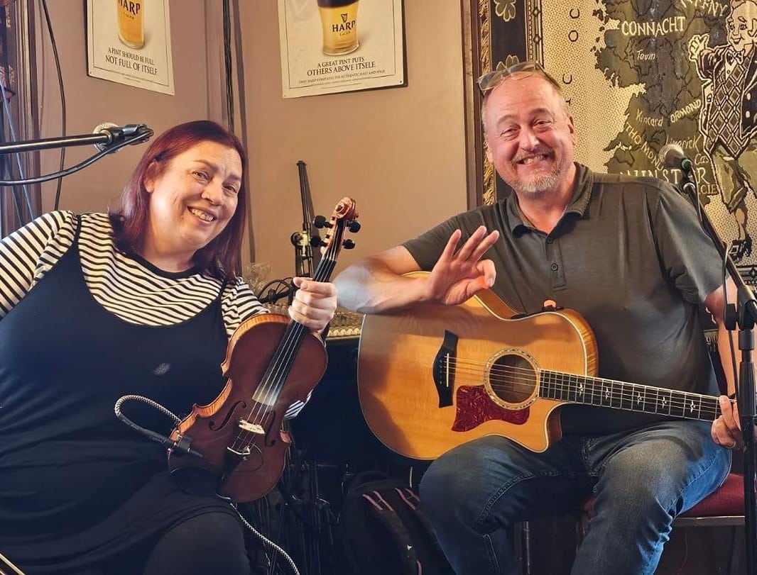 A woman holding a fiddle and a man holding an acoustic guitar sit together and smile in a pub.