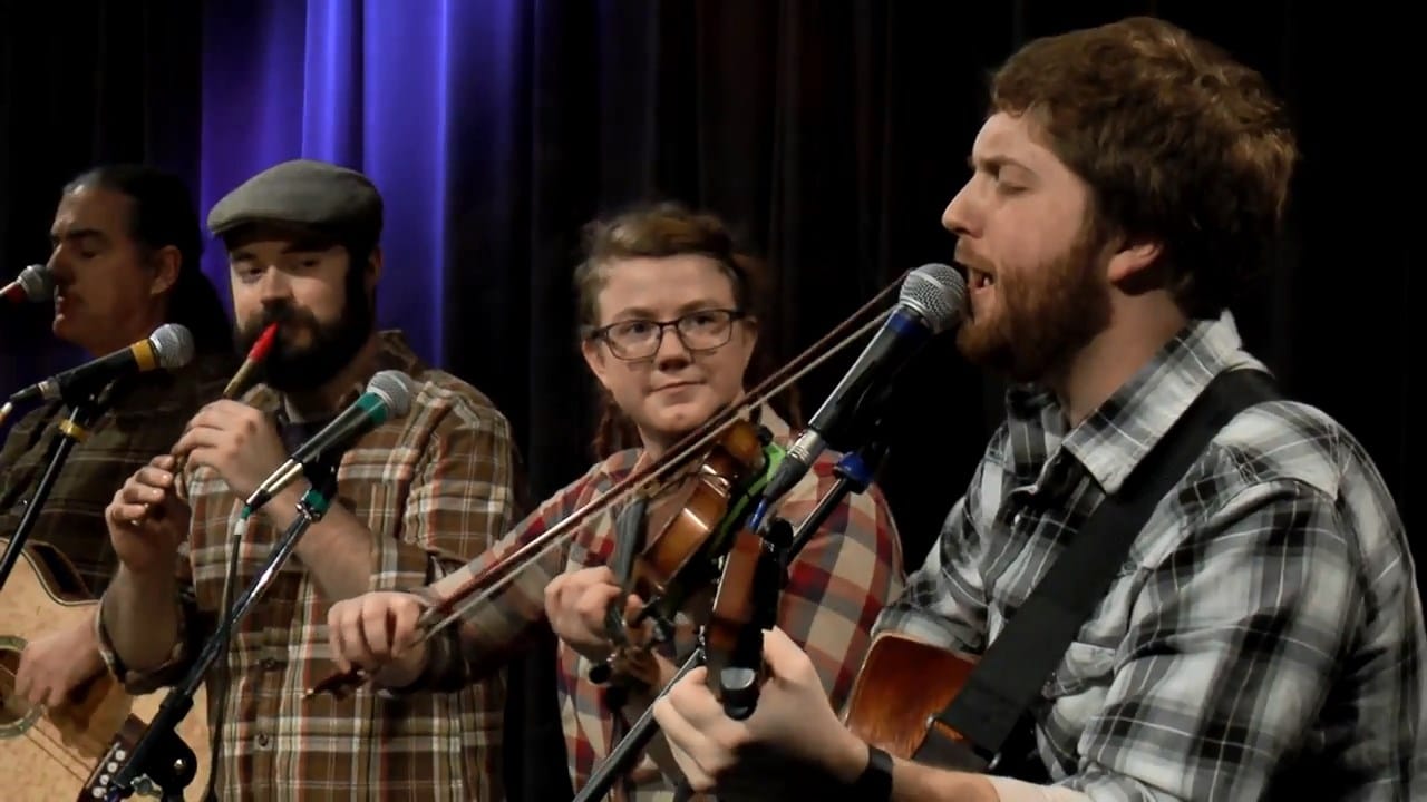 A band of four people playing acoustic instruments and singing into microphones on a stage with a dark curtain backdrop.