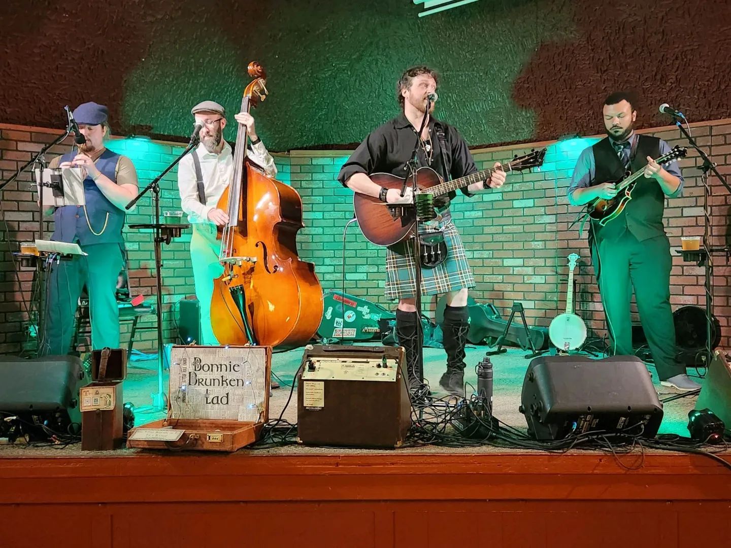 A four-piece folk band performs on stage with an upright bass, guitar, mandolin, and tin whistle.
