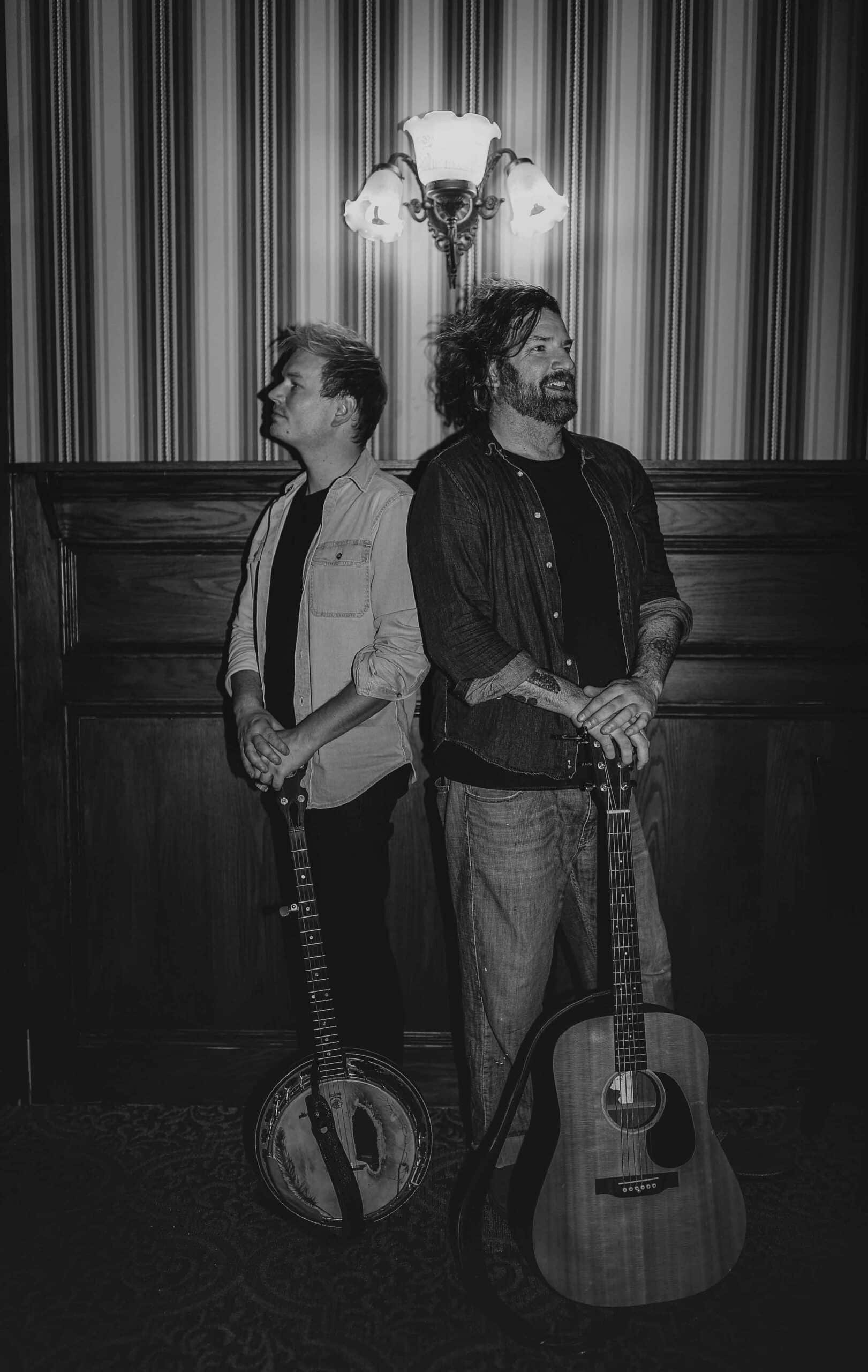 Two men, one holding a banjo and one holding an acoustic guitar, stand before a wood-paneled wall. Black and white.