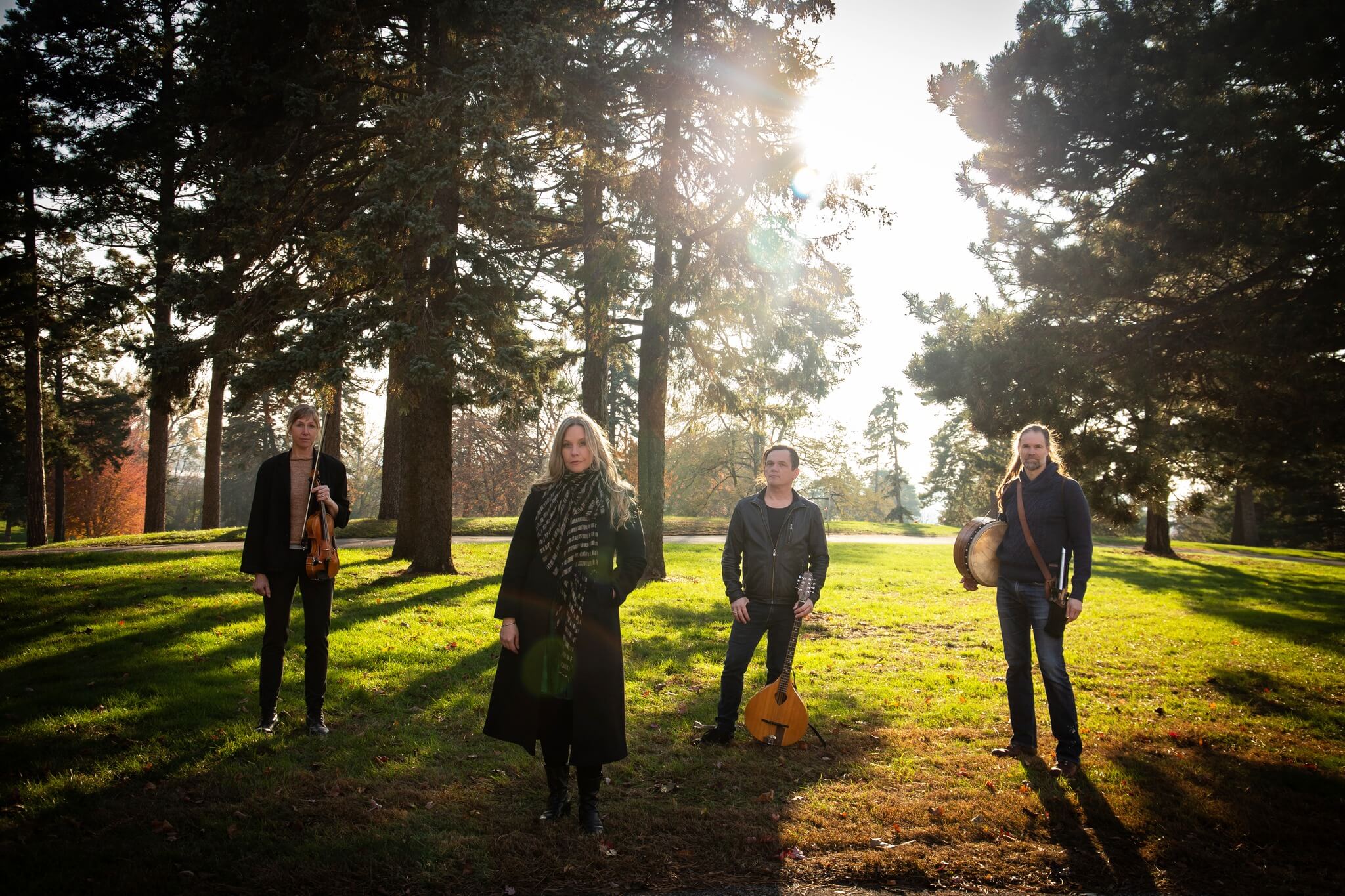Four musicians stand in a park with tall trees, backlit by the sun, holding their instruments.