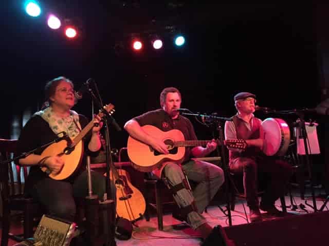 A musical trio plays acoustic instruments on a dark stage with spotlights overhead.