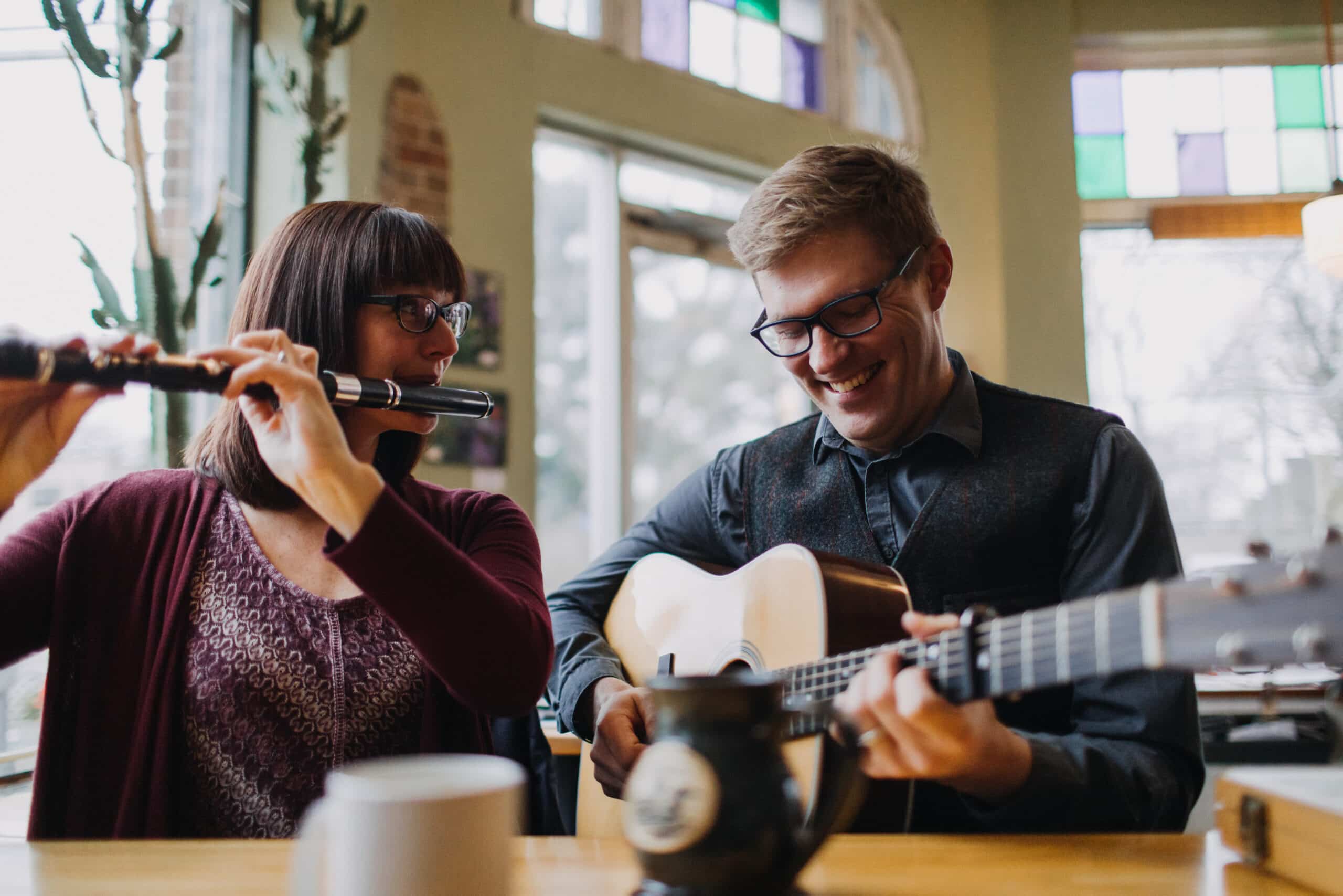 A woman plays the flute while a man plays the acoustic guitar in a cozy room with soft lighting.