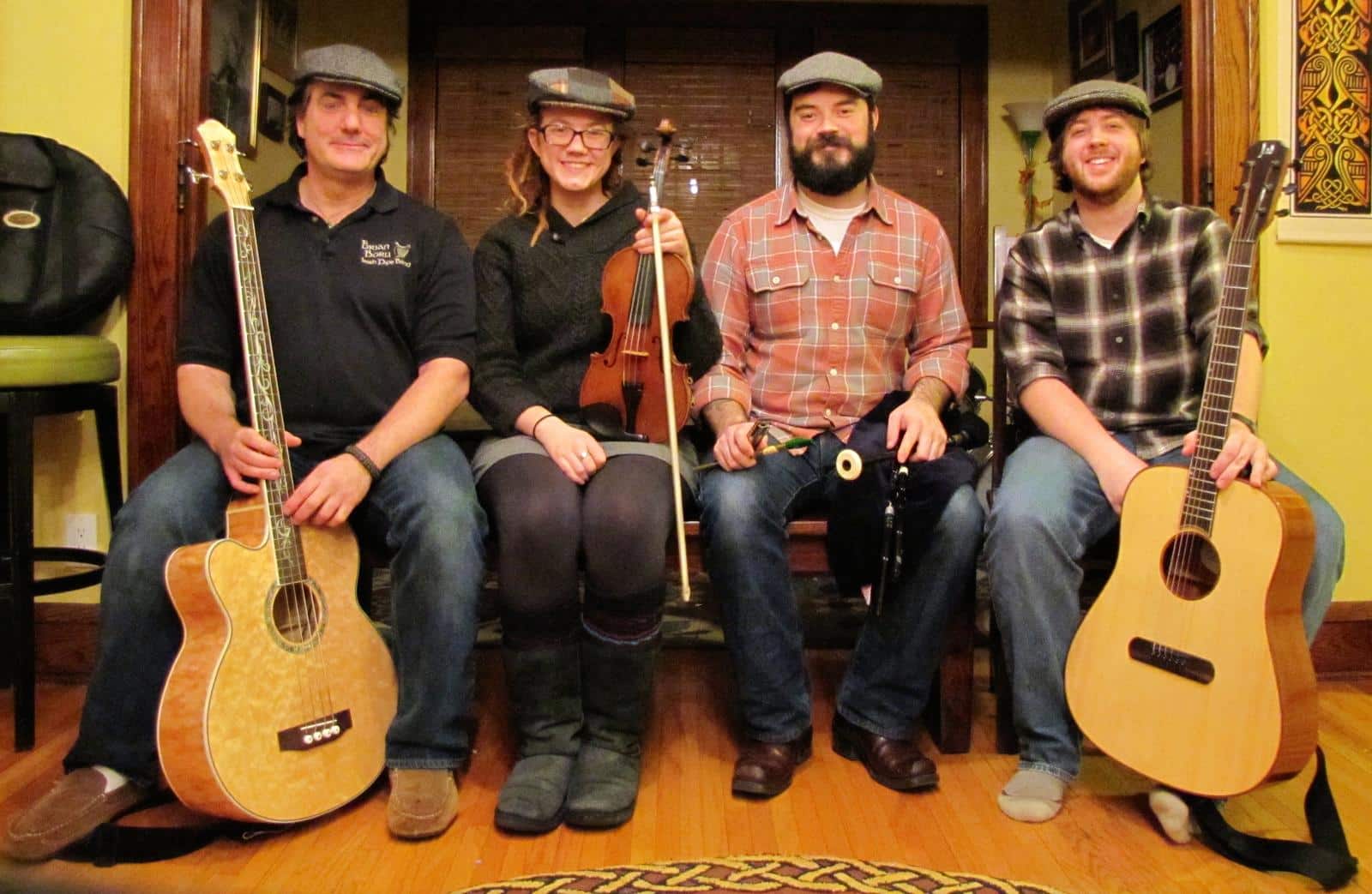 Four people in flat caps sit holding musical instruments, including guitars and a violin, in an indoor setting.
