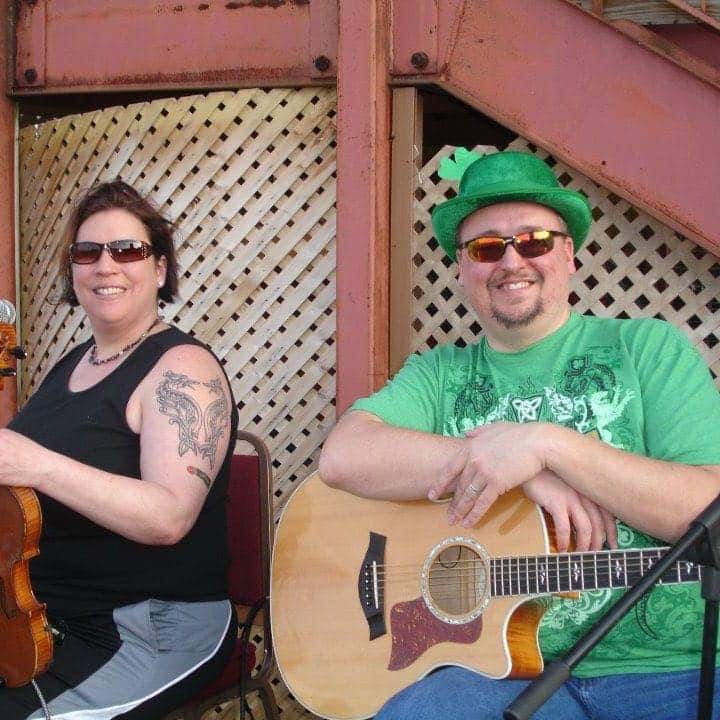 A woman playing a violin and a man in a green hat playing a guitar, both wearing sunglasses in front of a lattice wall.