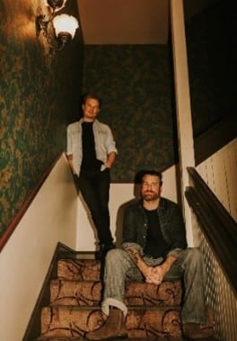 Two men pose on a carpeted staircase in a dimly lit hallway with patterned wallpaper and a wall lamp.
