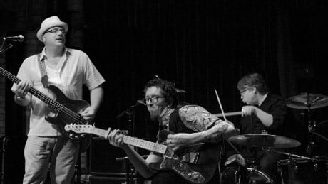 Black-and-white image of a musical trio performing on stage: a bassist, a guitarist, and a drummer.
