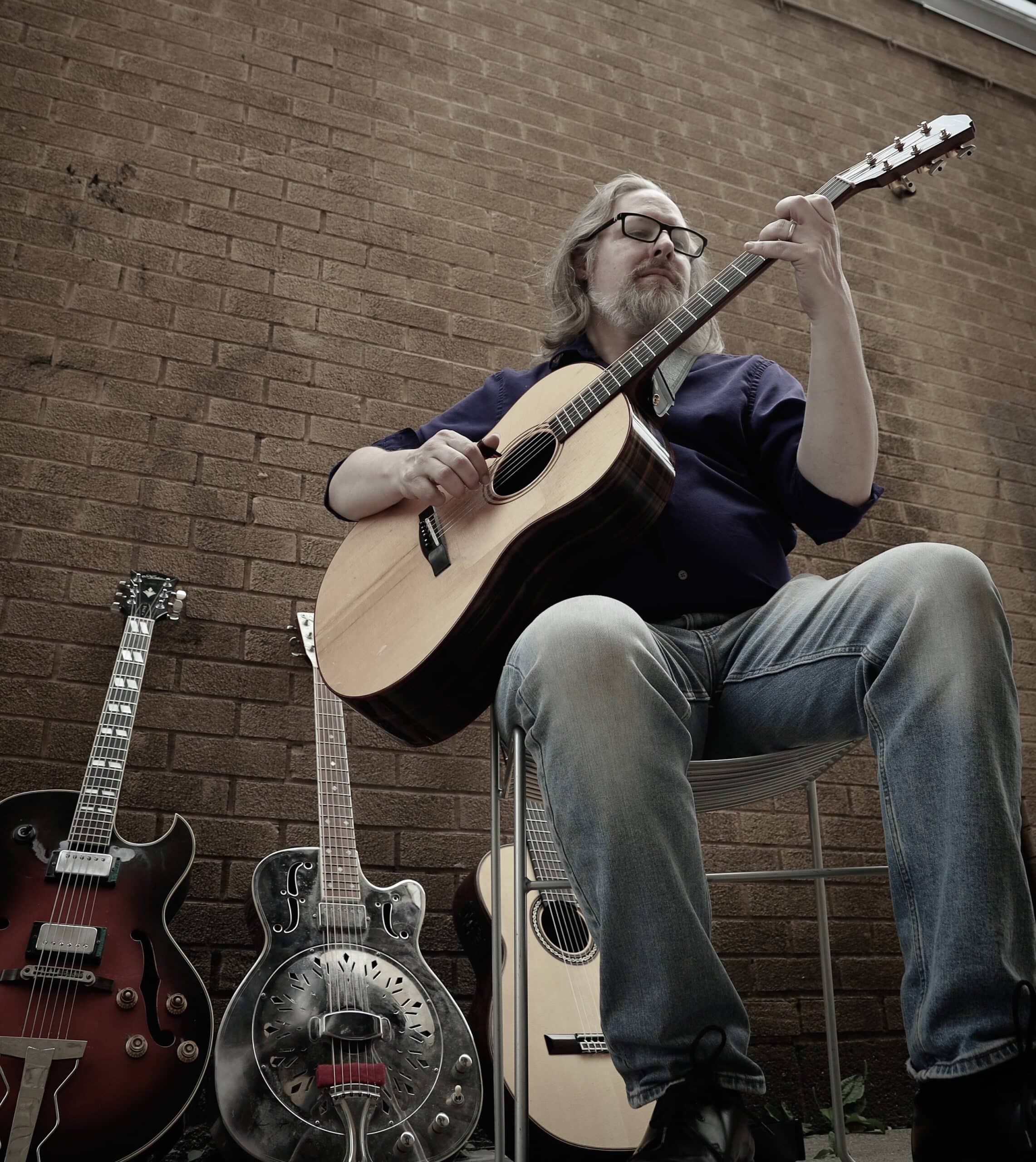 A man with long hair and glasses plays an acoustic guitar while sitting against a brick wall with three other guitars.