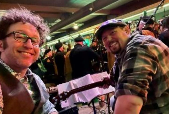 Two men smiling while holding guitars in a dimly lit venue with other people in the background.