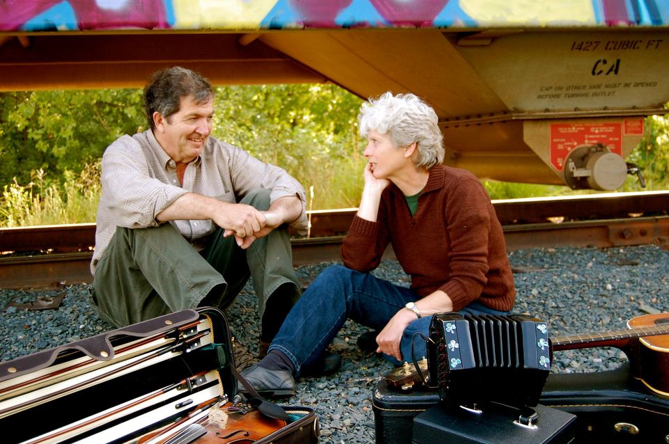 A man and woman sit on railroad tracks, facing each other and talking, with musical instruments on the ground.