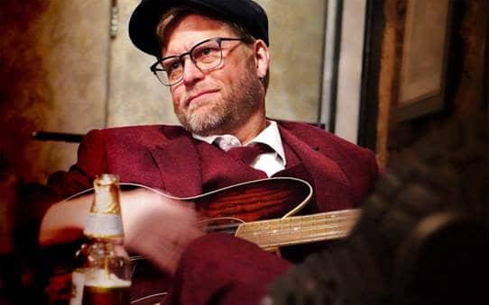 A man in a red suit, tie, and cap with glasses plays an acoustic guitar near a bottle of beer.