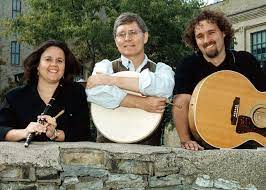 Three musicians pose behind a stone wall holding a flute, a bodhrán, and an acoustic guitar.