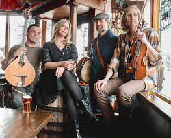 Four musicians sit in a rustic bar holding various string instruments, including a mandolin, banjo, and violin.