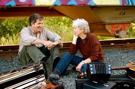 An older man and woman sit on railroad tracks facing each other, surrounded by musical instrument cases.