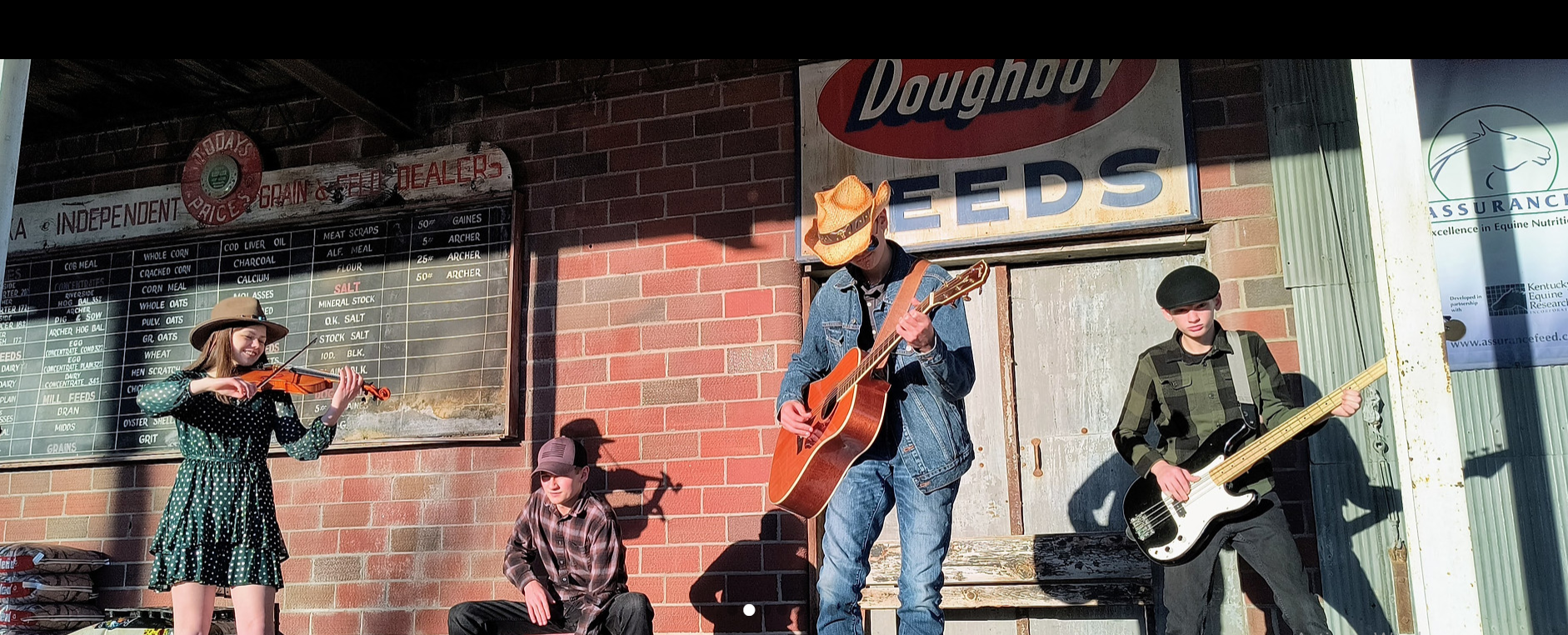 A band performs in front of a brick building with a Doughboy Seeds sign; one sits, three stand with instruments.