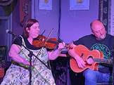 A woman plays the violin while a man plays an acoustic guitar on a stage under purple lighting.