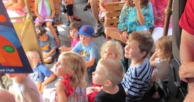 A group of children sits together, looking toward an off-screen storybook or presentation in a group setting.