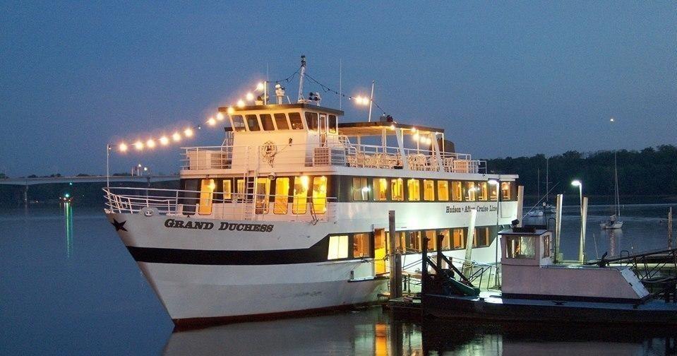 A white riverboat, named Grand Duchess, is docked on calm water at twilight, illuminated by festive string lights.