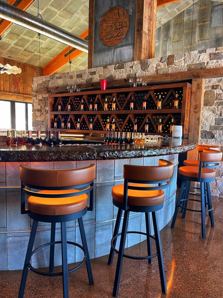 A bar with stone walls features three tan leather stools, a wine rack display, and a rustic wooden beam ceiling.