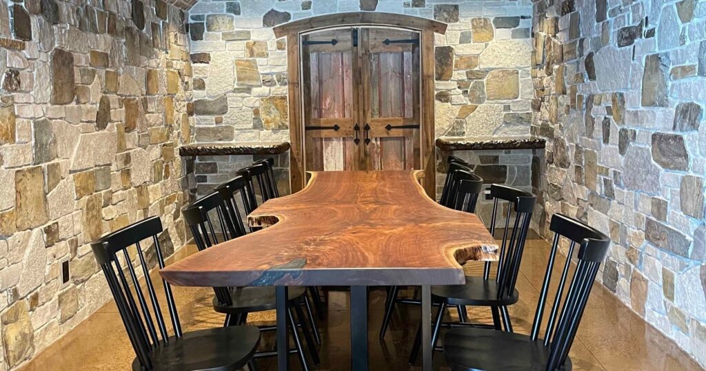 A large wooden live-edge table sits in a room with stone walls, black chairs, and a rustic wooden double door.