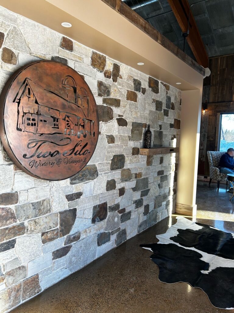 A stone wall features a round metal sign reading "Two Sile Winery & Vineyard," with a cowhide rug on the floor.