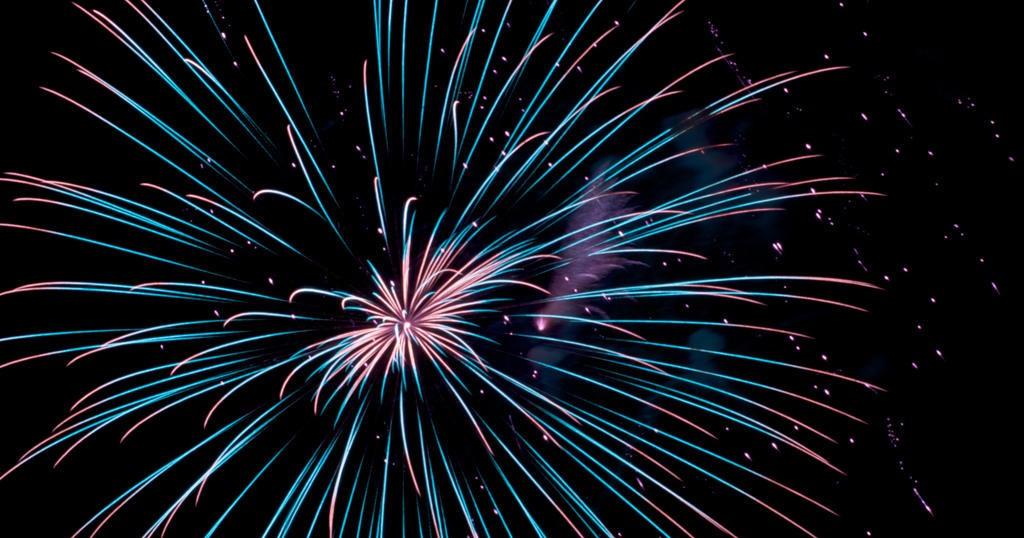 A vibrant firework display bursts against a dark night sky, with glowing blue and pink light streaks radiating outward.