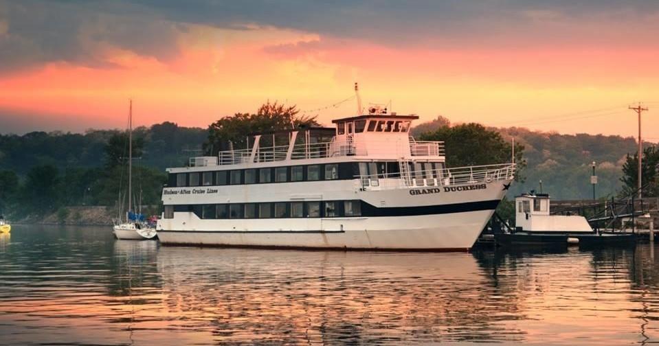 A white multi-deck tour boat sits on calm water at sunset, with a smaller vessel nearby against a hazy shore.
