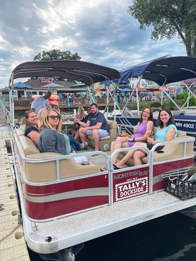 A group of people sit on a Tally's Dockside pontoon boat docked at a pier under a cloudy sky.