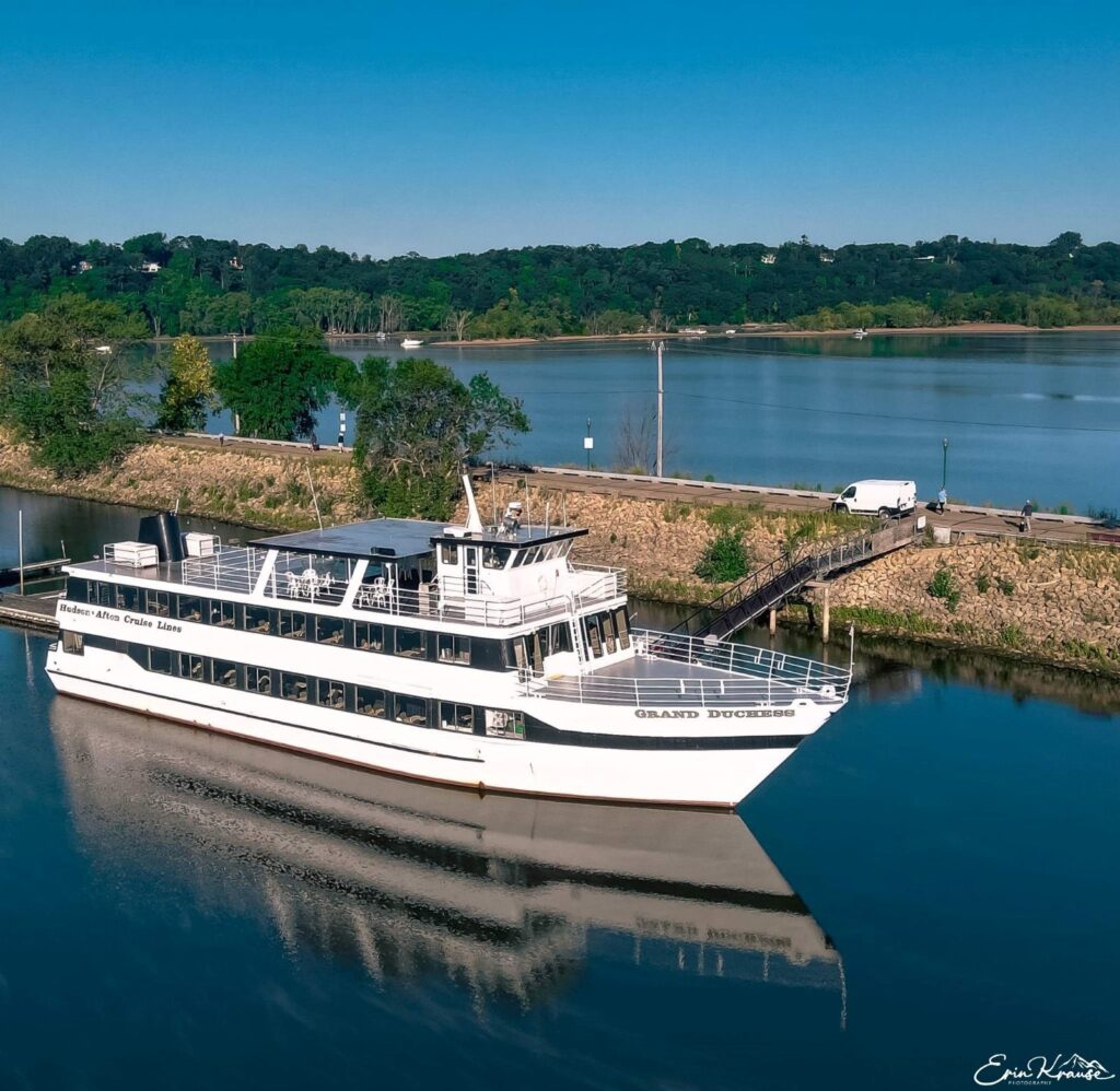 Aerial view of a white multi-deck passenger boat docked in a calm river near a grassy shore under a clear blue sky.
