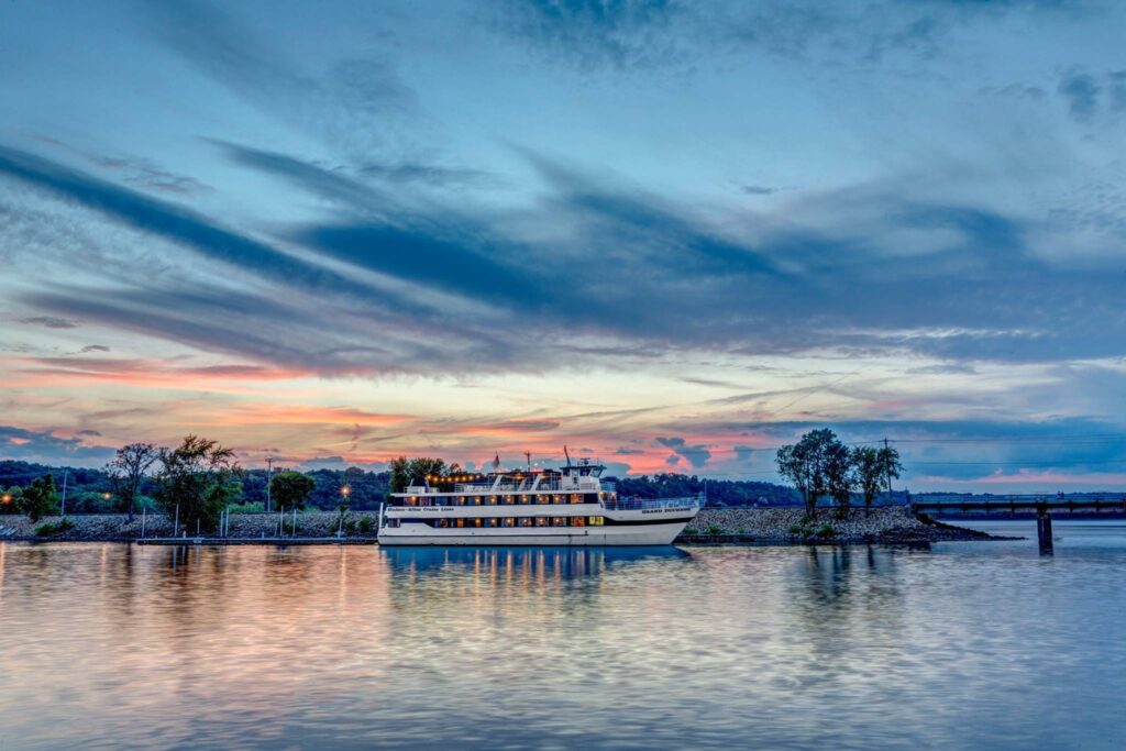 A passenger riverboat is docked on a calm river under a dramatic sunset sky with streaky clouds.