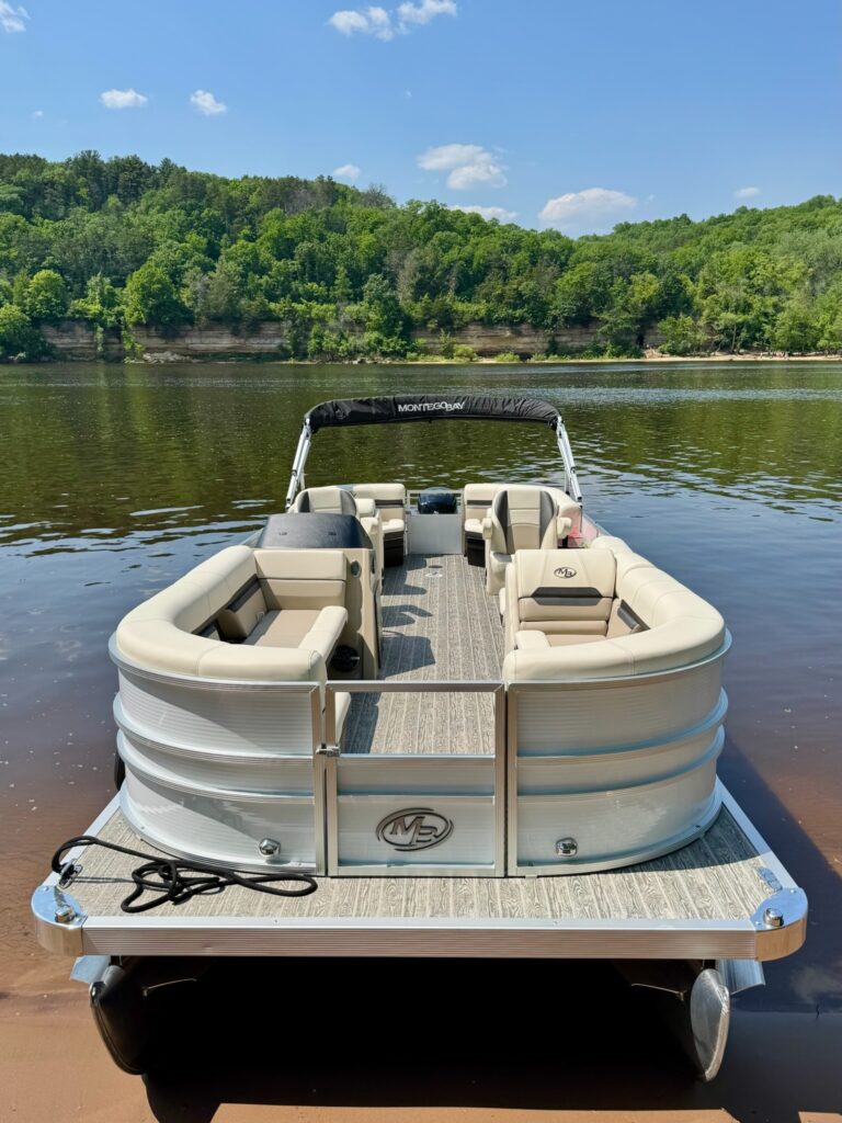 A view from the back of a pontoon boat docked on calm water with forested hills in the background under a blue sky.