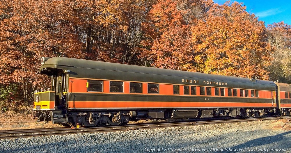 A Great Northern passenger train car parked on tracks in front of a forest with colorful autumn leaves.