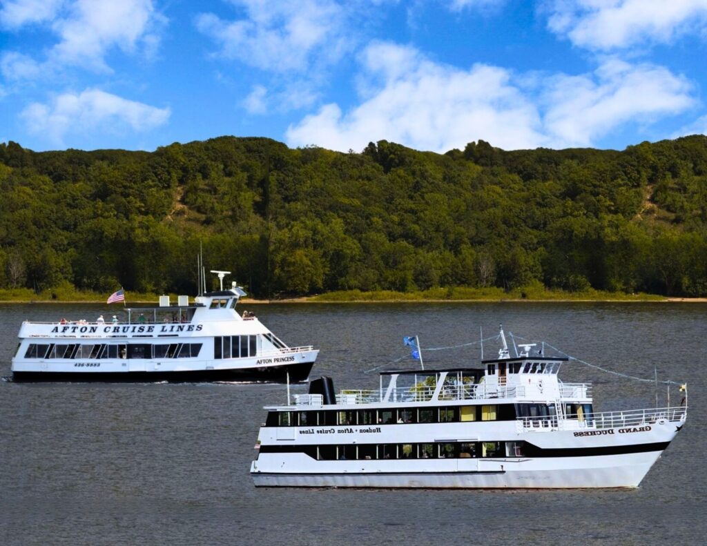 Two white cruise boats travel on a river in front of a tree-covered hill under a blue sky with clouds.