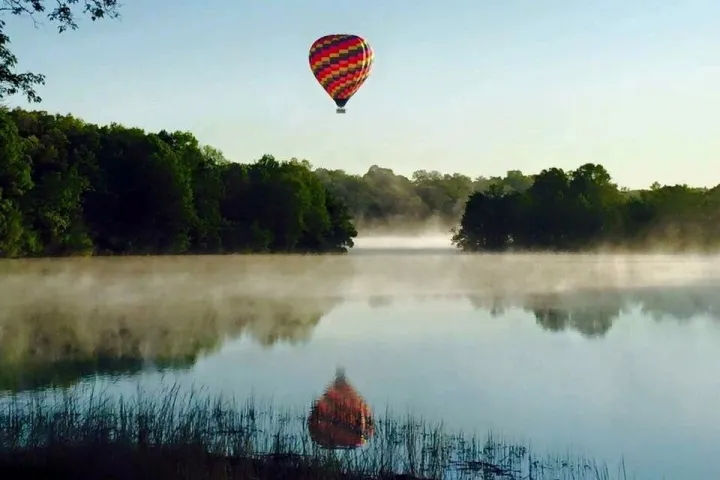 A hot air balloon floats above a misty lake, with trees on the shore and its reflection visible in the calm water.