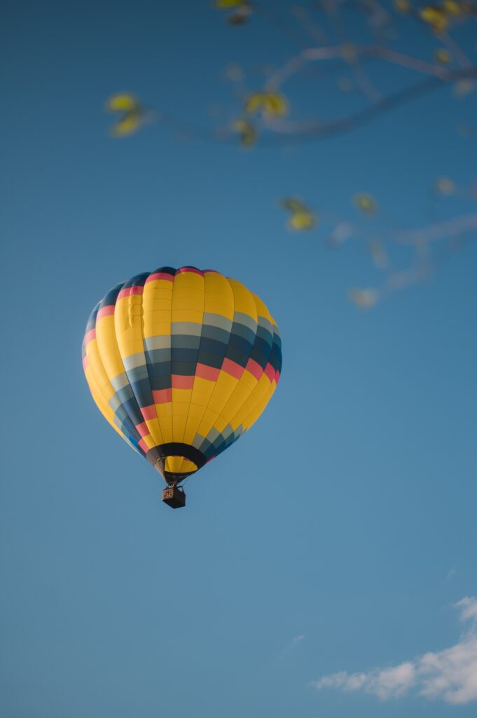 A yellow, blue, and orange patterned hot air balloon floats in a clear blue sky, with out-of-focus tree branches.