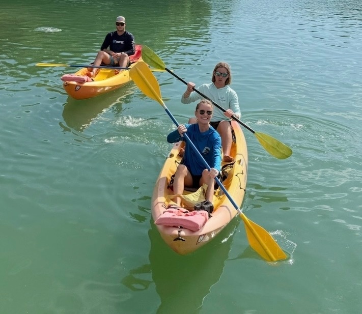 Three people in two kayaks are paddling on calm, green water.
