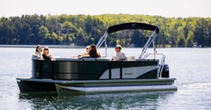 People riding a modern gray and black pontoon boat on a lake with a forest in the background.