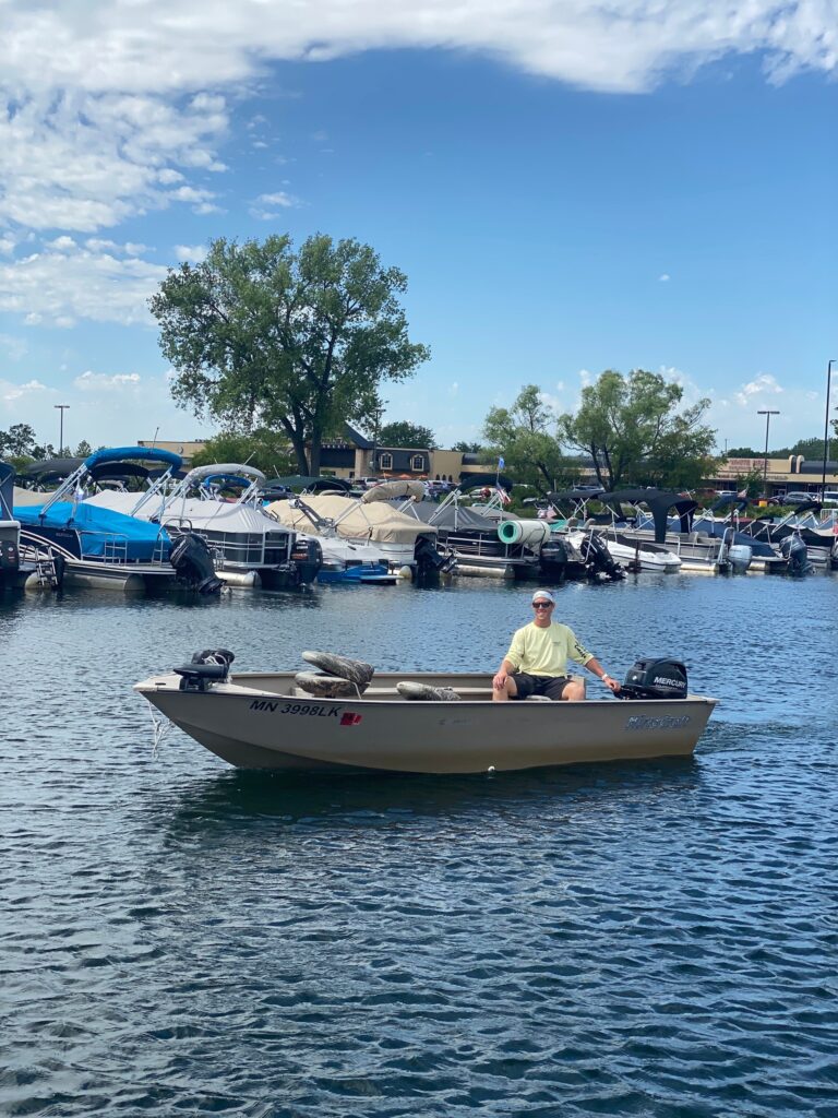 A person sits in a tan motorboat on a sunny day in a marina filled with other boats.