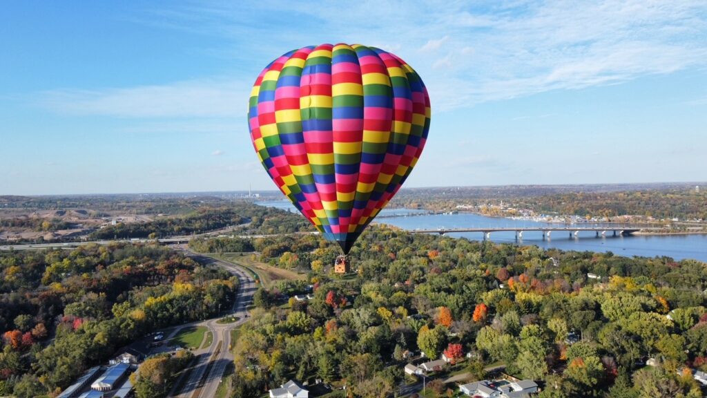 A colorful, multi-colored hot air balloon floats above a wooded landscape, river, and bridge.