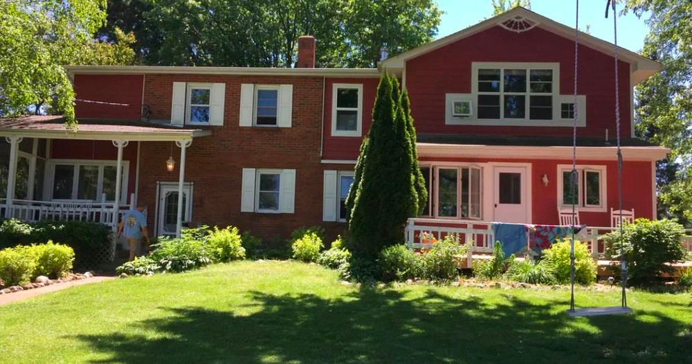 A large red house with a brick section, white shutters, and a front porch, viewed from a sunny lawn.