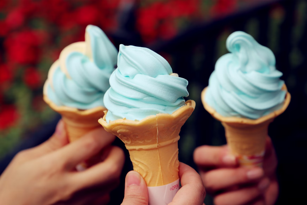 Hands hold three blue soft-serve ice cream cones in front of a blurry background of red flowers.