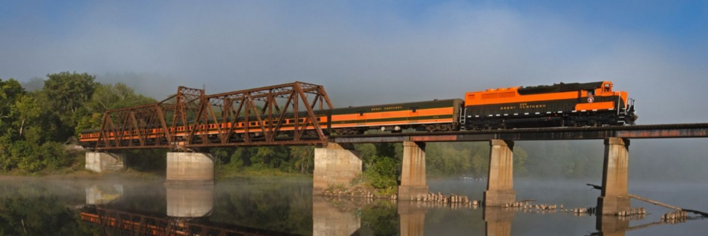 An orange and black train crosses a rusted truss bridge over a misty river at dawn.