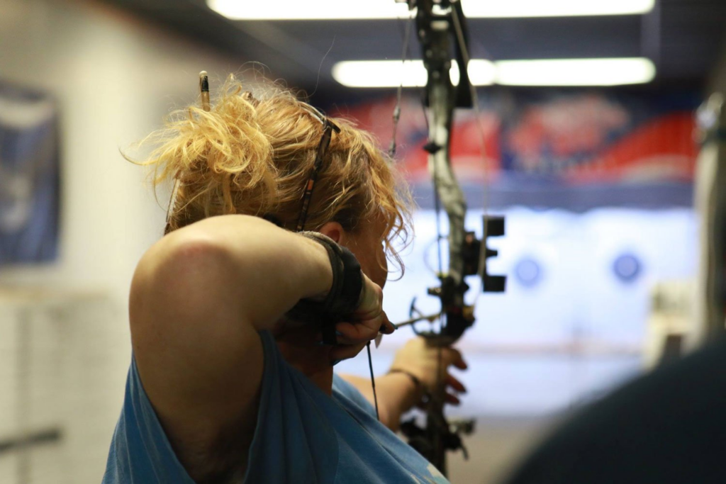 A woman with blonde hair holds a compound bow and aims at an indoor archery target.
