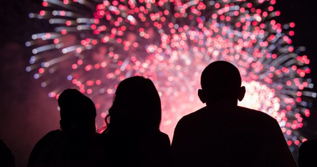 Silhouettes of three people watching a vibrant display of pink and white fireworks in the night sky.