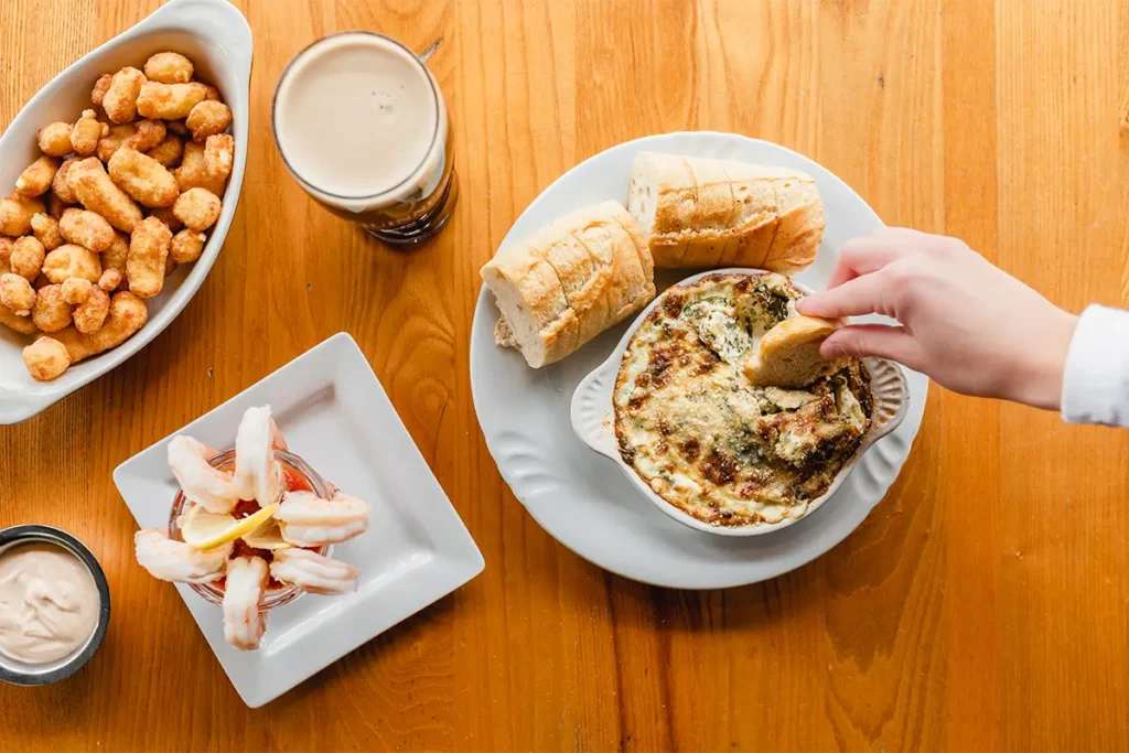 Top-down view of a wooden table with appetizers including cheese dip, bread, fried dough, shrimp, and a drink.