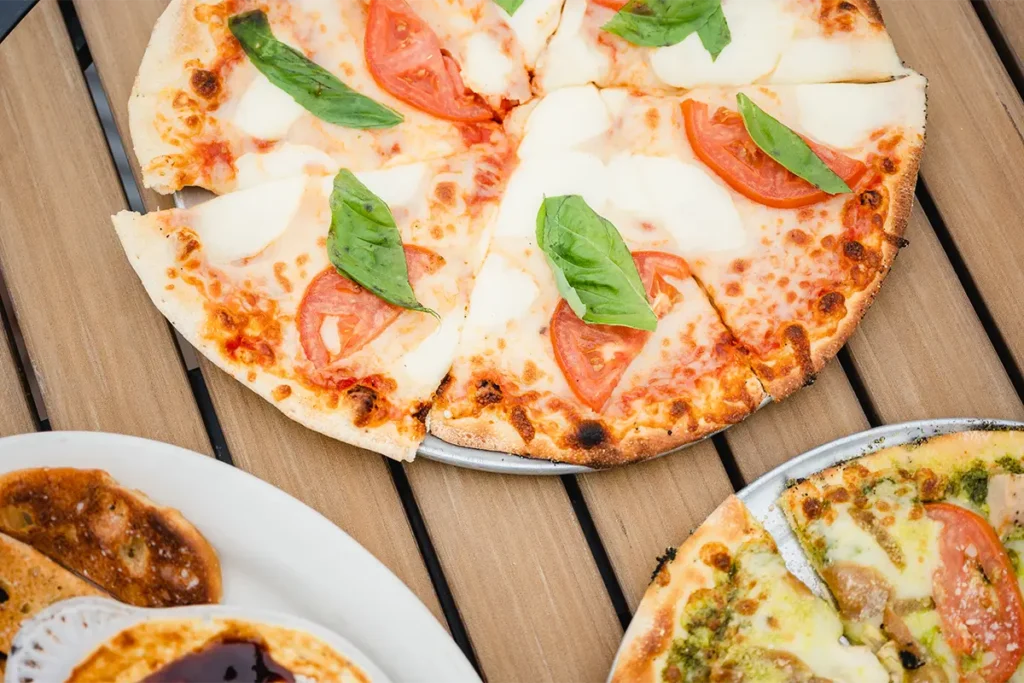 A Margherita pizza with tomatoes and basil sits on a wooden table, with other plates visible nearby.